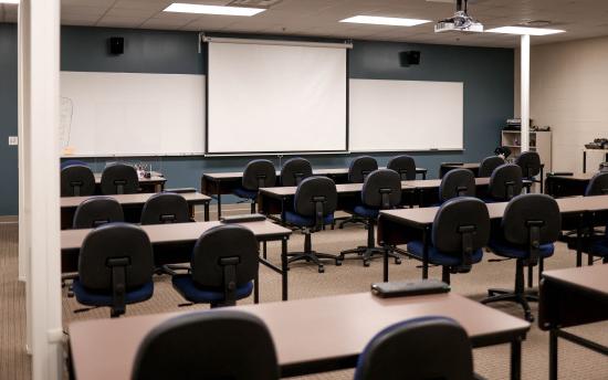 A traditional-style classroom with several tables and chairs with three projector screens in the front of the room